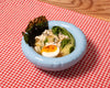 A round, baby blue ceramic bowl sits on top of a table. on the table, underneath the bowl is a red and white tablecloth. In the bowl is a ramen soup.