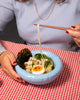 Ljósblá, kringlótt keramíkskál á borði. Undir skálinni á borðinu er köflóttur dúkur. Í skálinni er ramen súpa.