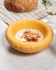 Kringlótt gul keramík skál á dúk lögðu borði. Í skálinni er hummus.