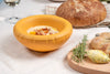 A round, yellow ceramic bowl on a table. In the bowl is hummus. A loaf of bread is next to the bowl on the table.