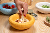 Three round ceramic bowls in various colours on a table. A baby blue coloured bowl with cherry tomatoes. A yellow bowl with hummus. A white bowl with pea salat.