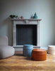 Three wonderful ceramic side- and coffee tables in beautiful colours in a livingroom. Wooden floors. A light grey lounge chair to the left. The side tables are light blue, earthy brown and white. In the background is a pale blue wall with a large fireplace.