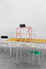 Two minimalistic dining tables. A small table with ivory coloured table top in the foreground while a larger table with a bright yellow table top is behind it. On each side is a colourful dining chair. A dining chair is on top of the smaller table while a red minimalistic lounge chair is on top of the larger one. Light grey concrete floor. A white wall in the background.