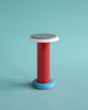 A pretty, multi coloured table lamp on a turquoise background. The lamp is red, white and blue with a black top.