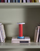 Pretty and colourful little lamp in red, white and blue. The lamp stands on top of two books in a book shelf.