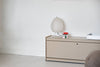 Plié Floor unit. Here in beige with one single drawer. It is made from powder coated aluminium. On it is a small table lamp. White wall in the background. White floor.