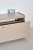 A close up of a beige coloured storage unit. Plié floor unit. It has one large drawer which is open in this photograph. On the floor unit is a small picture in black and white. White wall in the background. White floor in the foreground.