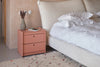 Beautiful pink side table with two drawers. Here in a master bedroom and used as a nightstand. on it is a small book and a small grey vase.