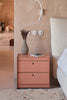 A small and wonderful pink side table with two drawers. It is made from powder coated aluminium. Here used as a nightstand and is next to a bed. On the nightstand is a small book and a small, grey vase. A white rug in the foreground. Behind the side table is a beige coloured wall with white light switches and a white reading light.
