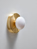 A gorgeous little wall sconce in soft shapes made from brushed brass and handmade, white glass. The sconce is mounted on a white wall. The sconce is turned off.