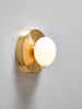 A gorgeous little wall sconce in soft shapes made from brushed brass and handmade, white glass. The sconce is mounted on a white wall. The sconce is turned on.