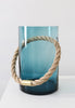 Large handmade glass vase in blue tinted colours. A pale coloured rope goes through the vase. The vase is placed on a white surface. White wall in the background.