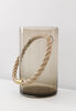 A gorgeous, light brown vase. Handmade glass. A beautifully woven roap goes through the vase. White wall in the background. The vase placed on a white surface 