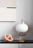 A gorgeous handmade glass sculpture in pink. The glass sculpture sits on top of a counter next to a table lamp in brass and white. White wall in the background. A white artwork hangs on the wall.