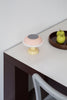Pale pink and yellow, cute little table lamp sits on the corner of a desk. White wall in the background. The lamp is turned off.