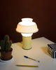 A very pretty little table lamp sits on a white table. The lamp is in pastel yellow colour with a white silicone lamp shade. Light is switched on.
