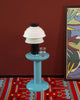 Beautiful, black table lamp with white silicone shade sits on top of a baby blue side table. Burgundy wall in the background. A very colourful rug on the floor. The light is switched off.