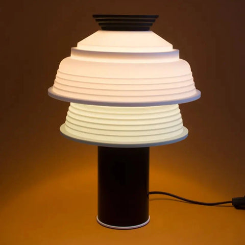 A striking little table lamp in black. It has a double lamp shade in white and mint green silicone. Brown background. The light is switched on.