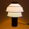 A striking little table lamp in black. It has a double lamp shade in white and mint green silicone. Brown background. The light is switched on.