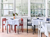 Interior image from a bright and beautiful restaurant. Red and black dining chairs nex to tables. Wooden floors. White walls with windows in the background.