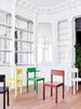 Interior shot from an old and beautiful building. Narrow shot. Five dining chairs are in the space. From left: a detail of a black chair. Bright yellow chair without arm rests. Bright green dining chair with arm rests. Red dining chair without arm rests and finally to the right; a white dining chair without arm rests.