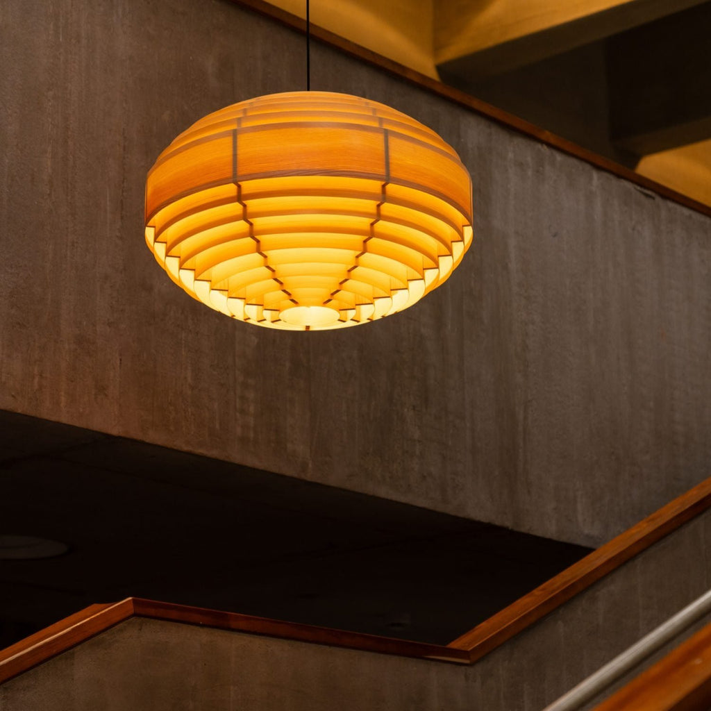 A large, spherical shaped pendant light made from pine veneer. Classic design by Hans-Agne Jakobsson. The light hangs above a staircase in a modernistic concrete building. The light is switched on.