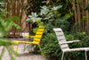 Two outdoor chairs in a beautiful garden on a summer day. The chair on the left is a yellow lounge chair and the one on the right is a white dining chair with armrests. Lots of trees and flora.