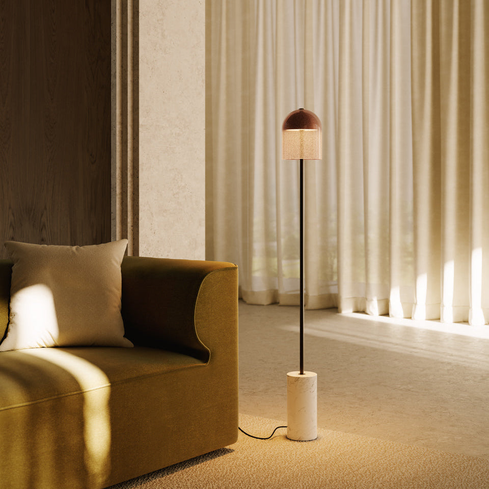 Fágaður, grannur gólflampi frá Venicem. Base úr hvítum marmara. Dökk brennt brass og handgert gler. Lampinn stendur í stofu. Ljós beige litapalletta. Gluggatjöld fyrir glugga í bakgrunni. Kveikt er á lampanum.