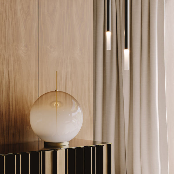 Afskaplega fágaður, kúlulaga borðlampi úr hvítu og gegnsæju gleri. Lampinn stendur á skenk. Viðarveggur í bakgrunni.