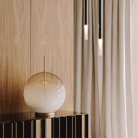 Afskaplega fágaður, kúlulaga borðlampi úr hvítu og gegnsæju gleri. Lampinn stendur á skenk. Viðarveggur í bakgrunni.