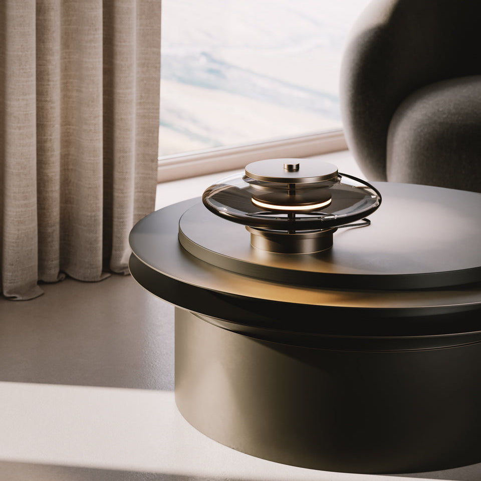 Fágaður borðlampi úr reyktu bronze. Lampinn stendur á sófaborði. Kveikt er á lampanum.