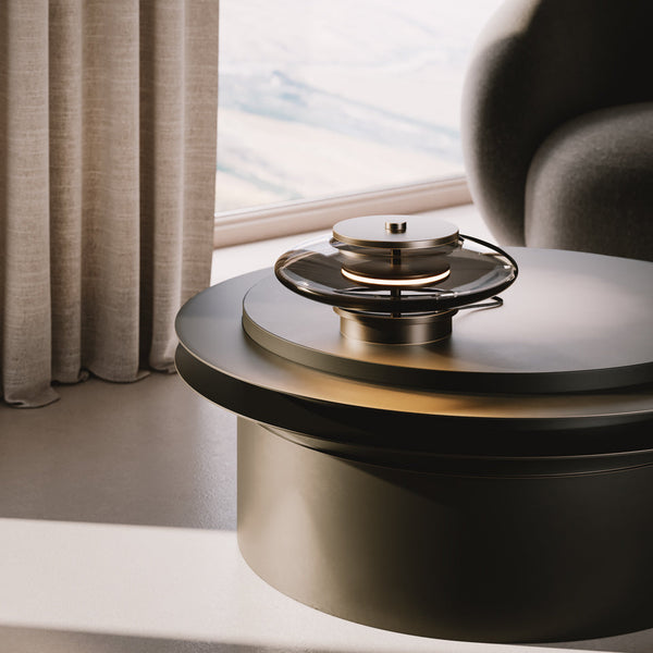 Fágaður borðlampi úr reyktu bronze. Lampinn stendur á sófaborði. Kveikt er á lampanum.