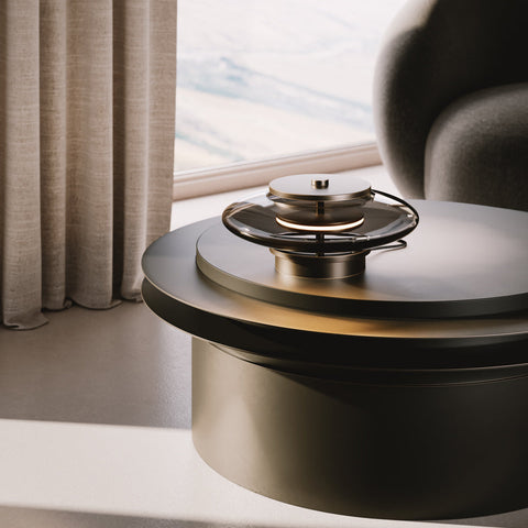 Fágaður borðlampi úr reyktu bronze. Lampinn stendur á sófaborði. Kveikt er á lampanum.