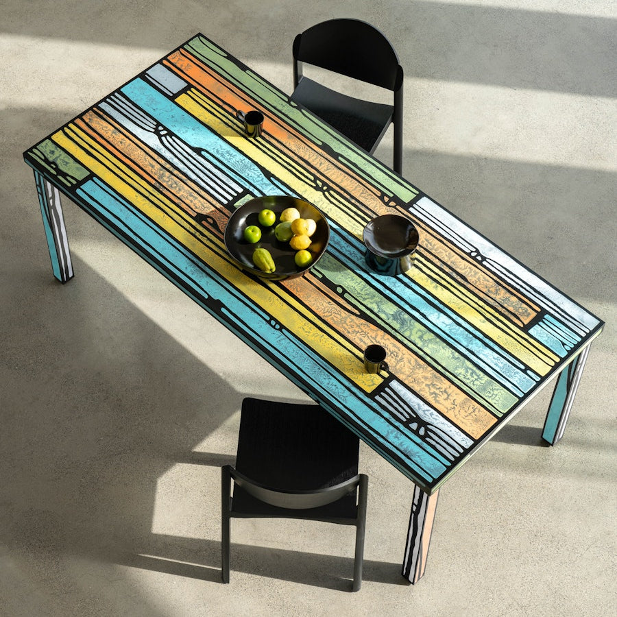 Afar litríkt borðstofuborð eftir Richard Woods og Sebastian Wrong á steyptu gólfi. Tveir svartir borðstofustólar við borðið. Ávaxtaskál á borðinu. Sjónarhorn að ofanverðu.