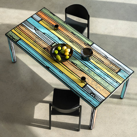 Afar litríkt borðstofuborð eftir Richard Woods og Sebastian Wrong á steyptu gólfi. Tveir svartir borðstofustólar við borðið. Ávaxtaskál á borðinu. Sjónarhorn að ofanverðu.