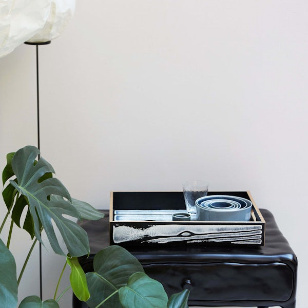 Fallegur og listrænn bakki í hvítum og svörtum lit eftir Richard Woods og Sebastian Wrong situr á svörtu náttborði. Hvítur veggur í bakgrunni.