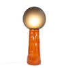A large ceramic floor lamp in earthy orange colour. Handmade glass spheric defuser in smokey grey. White background. The lamp is lit.