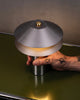 Hönd heldur utan um nettann borðlampa úr áli. Lampinn stendur á ólífugrænum skenk úr stáli. Kveikt er á lampanum.