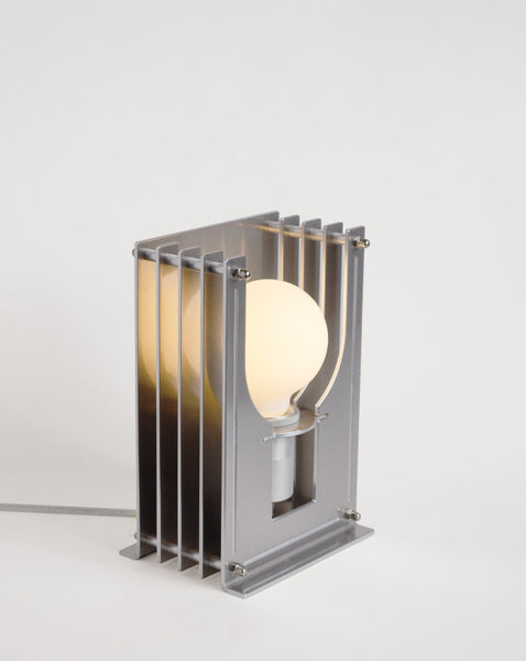 Silfurlitaður og skúlptúrískur borðlampi. Hvítur bakgrunnur. Kveikt er á lampanum.