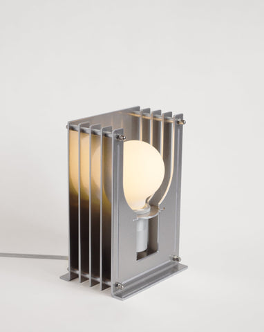 Silfurlitaður og skúlptúrískur borðlampi. Hvítur bakgrunnur. Kveikt er á lampanum.