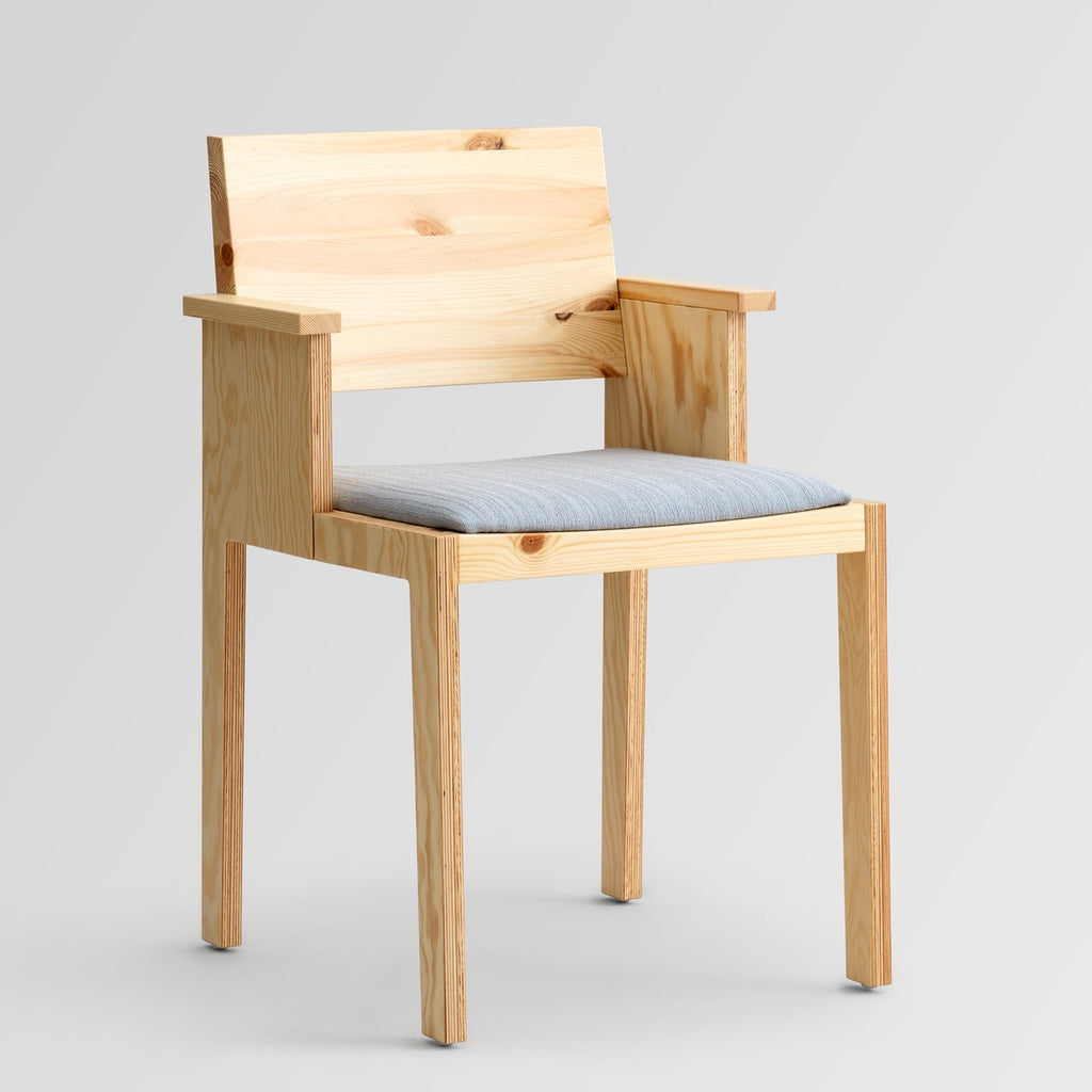 Borðstofustóll með örmum úr furu. Grátt áklæði á setu. Ljós grár bakgrunnur. Sjónarhorn á ská að framanverðu.