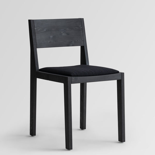 Svartur borðstofustóll án árma úr furu. Svart áklæði á setu. Ljós grár bakgrunnur. Sjónarhorn á ská að framanverðu.