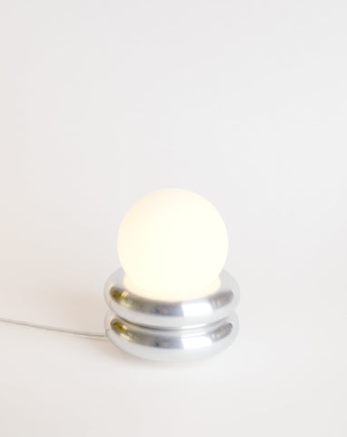 Sætur lítill, silfurlitaður borðlampi. Hvítur bakgrunnur. Kveikt er á lampanum.