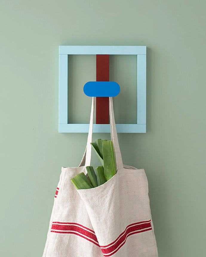 Fallegur, litríkur og glaðlegur veggsnagi eftir Nahalie Du Pasquier á ljósgrænum vegg. Hvítur taupoki með rauðri rönd hangir á snaganum.