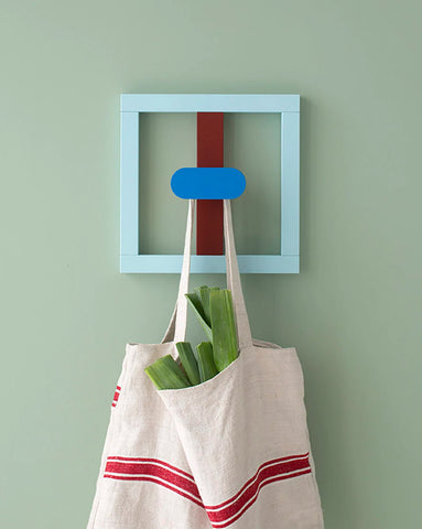 Fallegur, litríkur og glaðlegur veggsnagi eftir Nahalie Du Pasquier á ljósgrænum vegg. Hvítur taupoki með rauðri rönd hangir á snaganum.
