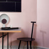 Narrow interior shot. A dark dining chair in the foreground. An end of dining table visible on the left. Light gray table lamp with an oval shape and black tray on the table. Bright pink wall in the background. Dark blue pin board on the wall.