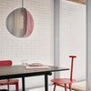 Pendant light in grey, pink and brass hangs above a black dining table. Two red dining chairs. White, brick wall in the background.