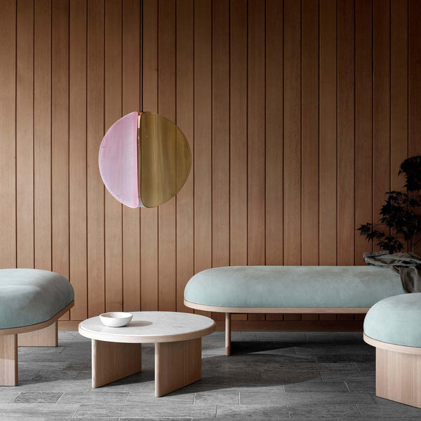 Pendant in pink and brass. Elegant benches and coffee table. Wooden wall in the background. Grey tiles on the floor.