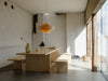 A large and gorgeous pendant light made from bendt pine veneer. The light is in a bright and beautiful dining room filled with sunlight. A pine dining table with benches. Light grey concrete floor. White walls in the background.