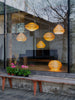 View from the outside through a large window. Bench outside in front of the window. On the inside of the house, behind the window hang five beautiful pendant lights made from bendt pine veneer in various shapes. Another light in the same style is on the floor. All of the lights are turned on.